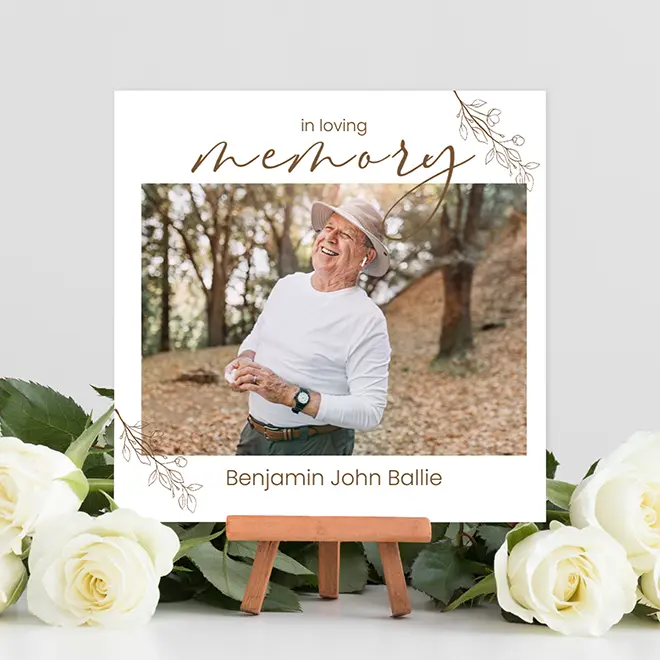 "Square contemporary memory photo print featuring a smiling elderly man in a white shirt and hat, set against an autumnal background. The print includes the text 'in loving memory' and a name, with delicate floral accents. Displayed on a small wooden easel, surrounded by white roses."