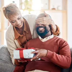 Image of a girl playfully blocking a man's eyes while surprising him with a gift, capturing a romantic and fun Valentine's Day moment, perfect for inspiring gift ideas.