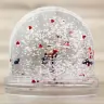 Clear plastic snow globe with a dome shape, filled with white snow-like particles and red heart-shaped confetti. The base is transparent, and inside the globe, there is a small figurine. Ideal as a personalised gift, suitable for occasions like Valentine's Day or Christmas.