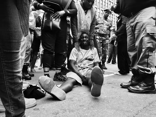 Black and white mobile photograph by Deen Schroeder, featuring a man on the floor with people standing around him