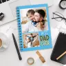 Spiral-bound personalised photo book with a blue cover featuring two photos of a father and child, and the text ''Best Dad Ever.'' Ideal for gifts for dad, birthday presents, or Father''s Day. The photo book is placed on a desk with office supplies, a coffee cup, and a laptop.