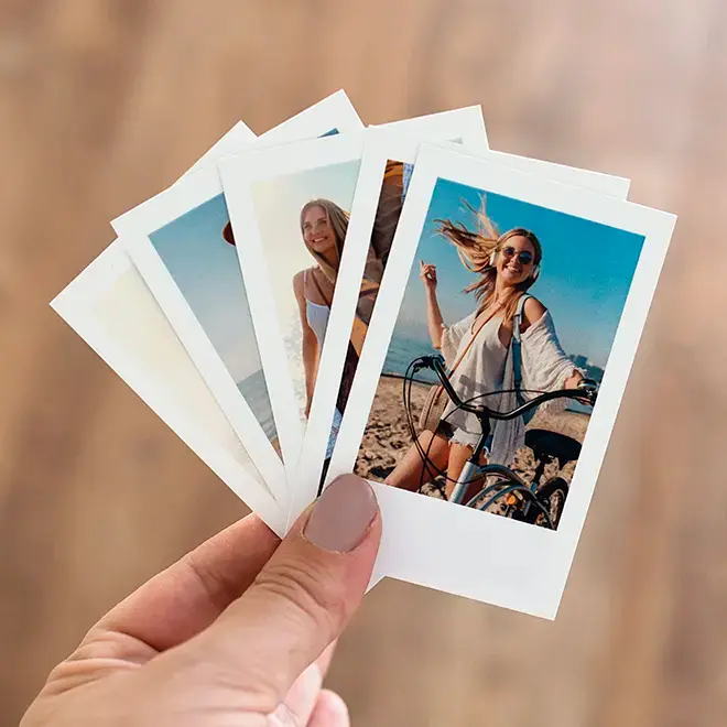 A hand holding a set of five printed photo cards, each featuring different outdoor scenes with people. The photos have a white border, resembling Polaroid-style prints. The background is blurred, highlighting the photo cards. Ideal for personalised gifts and photo albums.