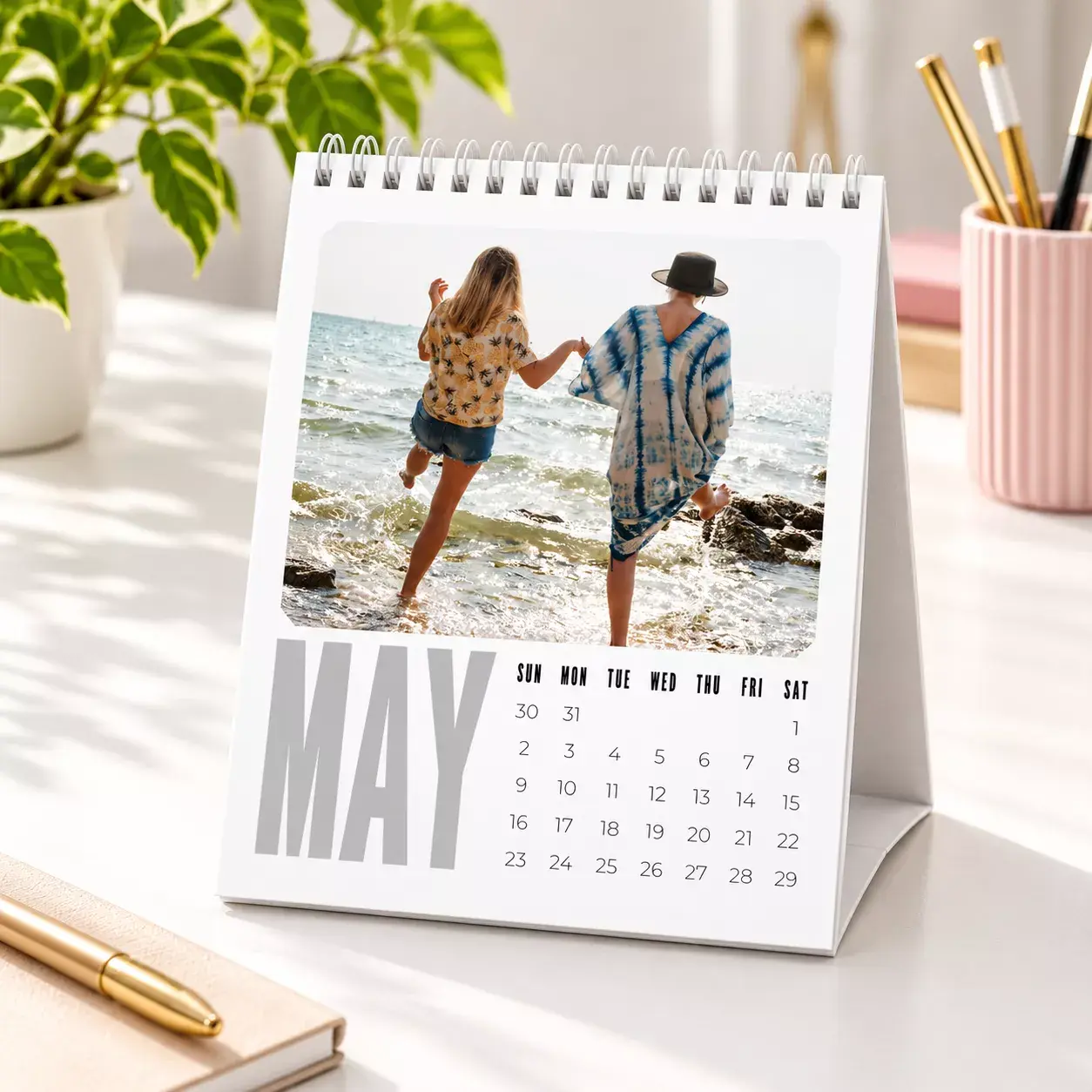 Personalised desk calendar featuring a photo of a baby in a colourful inflatable pool, wearing heart-shaped sunglasses and a pink hat. The calendar has a spiral binding and a stand, with the text 'Let's Make more Memories' at the bottom. Ideal for gifts and keepsakes.