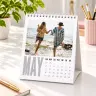 Personalised desk calendar featuring a photo of a baby in a colourful inflatable pool, wearing heart-shaped sunglasses and a pink hat. The calendar has a spiral binding and a stand, with the text 'Let's Make more Memories' at the bottom. Ideal for gifts and keepsakes.