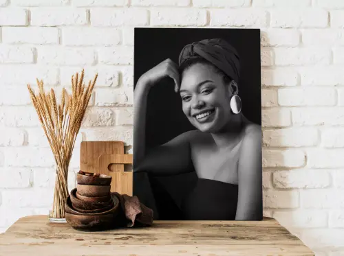 "Stylish black and white wall art featuring a smiling woman with a headwrap and large earrings, displayed against a white brick wall. The artwork is placed on a wooden surface adorned with rustic decor items, including wooden bowls and dried wheat stalks. Ideal for personalised gifts in South Africa, photo to canvas, and home decor."