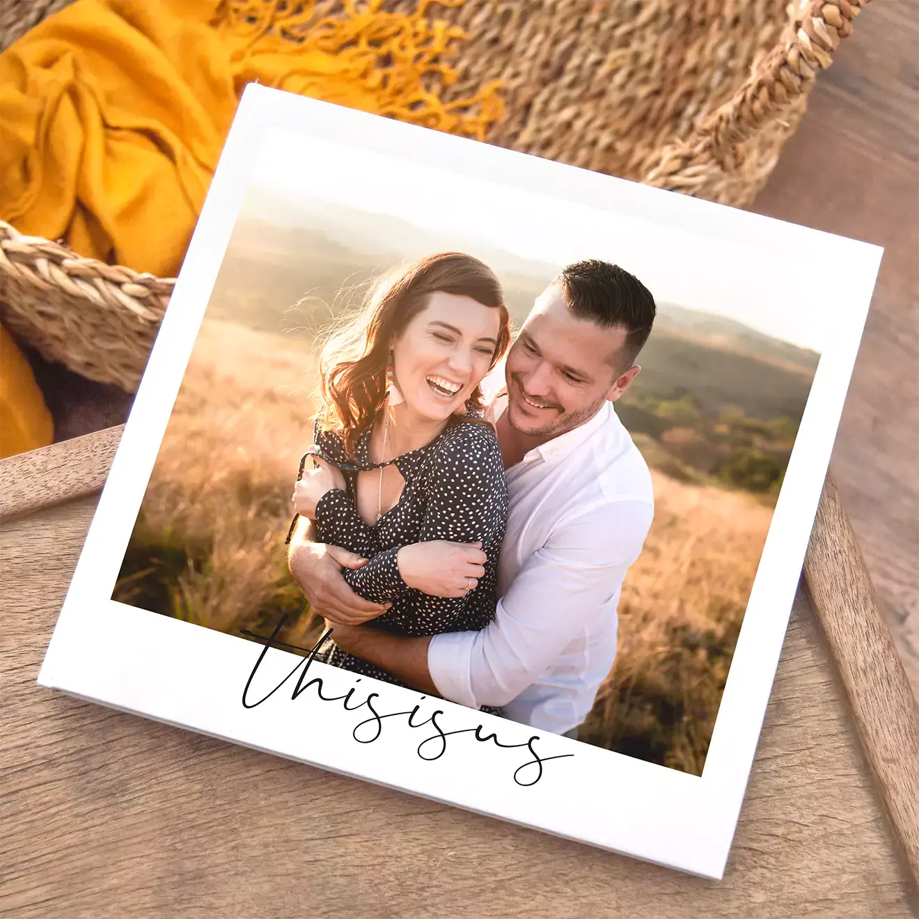 Square photo book with a white cover featuring a couple embracing in a scenic outdoor setting. The cover has the text "this is us" in elegant script at the bottom. The book is placed on a wooden surface with a woven basket and yellow fabric in the background. Ideal for personalised gifts in South Africa.