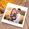 Square photo book with a white cover featuring a couple embracing in a scenic outdoor setting. The cover has the text "this is us" in elegant script at the bottom. The book is placed on a wooden surface with a woven basket and yellow fabric in the background. Ideal for personalised gifts in South Africa.
