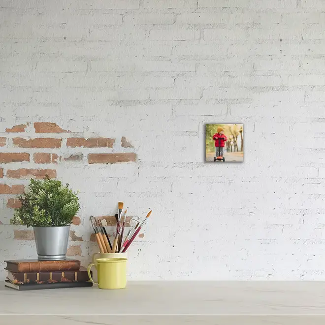 Small personalised canvas print featuring a child in a red coat, displayed on a white brick wall above a desk. The desk holds a potted plant, a stack of books, a yellow mug, and a jar of paintbrushes. Ideal for gifts for men, Christmas gift ideas, and personalised gifts in South Africa.