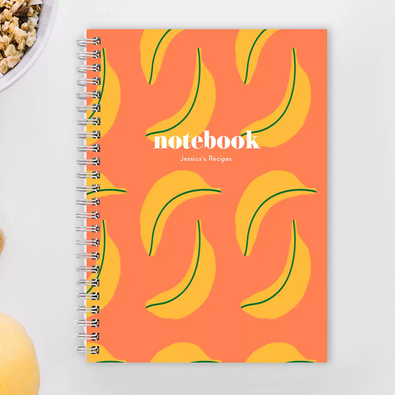 Spiral-bound notebook with a vibrant orange cover featuring a playful banana pattern. The word ''notebook'' is printed in white, with space for personalised text below. Ideal for recipe collections, journaling, or note-taking. Perfect as a gift for various occasions.