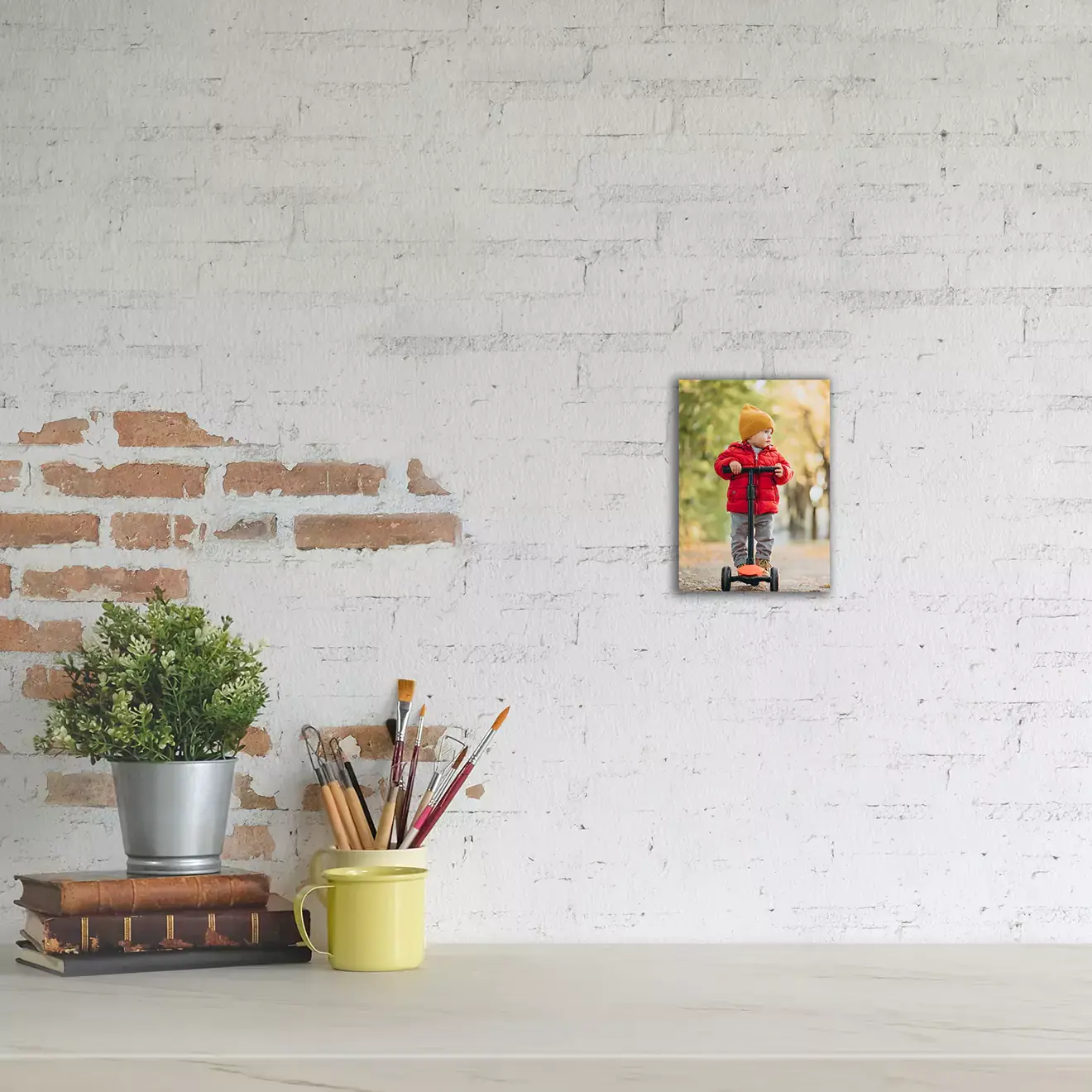 A small canvas photo print featuring a child in a red jacket and yellow hat riding a scooter, mounted on a white brick wall. The canvas is displayed above a desk with a potted plant, a stack of books, and a yellow mug holding paintbrushes. Ideal for personalised gifts in South Africa.