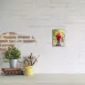 A small canvas photo print featuring a child in a red jacket and yellow hat riding a scooter, mounted on a white brick wall. The canvas is displayed above a desk with a potted plant, a stack of books, and a yellow mug holding paintbrushes. Ideal for personalised gifts in South Africa.