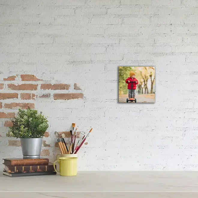 A small square photo canvas featuring a child in a red jacket riding a scooter in an autumn park, displayed on a white brick wall. The canvas is part of a minimalist decor setup with a potted plant, a stack of books, and a yellow mug holding paintbrushes on a white surface. Ideal for personalised gifts in South Africa.