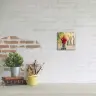 A small square photo canvas featuring a child in a red jacket riding a scooter in an autumn park, displayed on a white brick wall. The canvas is part of a minimalist decor setup with a potted plant, a stack of books, and a yellow mug holding paintbrushes on a white surface. Ideal for personalised gifts in South Africa.