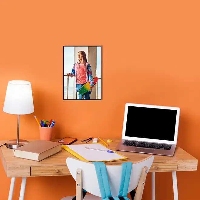 A stylish workspace featuring a wooden desk with a white chair, a laptop, a lamp, and stationery. The desk is set against a vibrant orange wall, adorned with a personalised A3 canvas photo print of a person holding a skateboard. Ideal for home office decor and personalised gifts in South Africa.