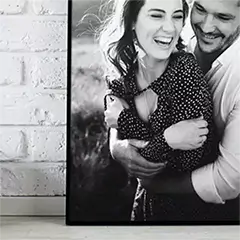 Contemporary framed canvas featuring a black and white photo of a smiling couple embracing. The canvas has a sleek black frame and is displayed against a white brick wall. Ideal for personalised gifts in South Africa, photo to canvas, and Christmas gift ideas.