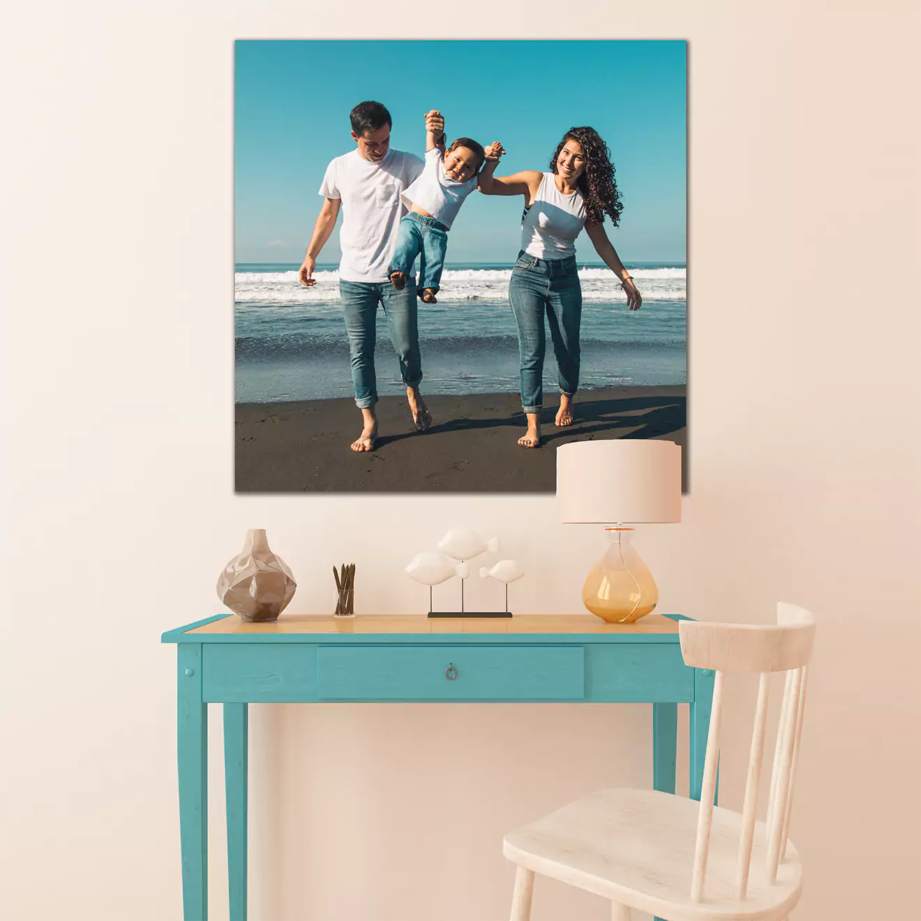 A family photo canvas print displayed above a turquoise desk with a white chair. The photo shows a family of three walking on the beach, holding hands and smiling.