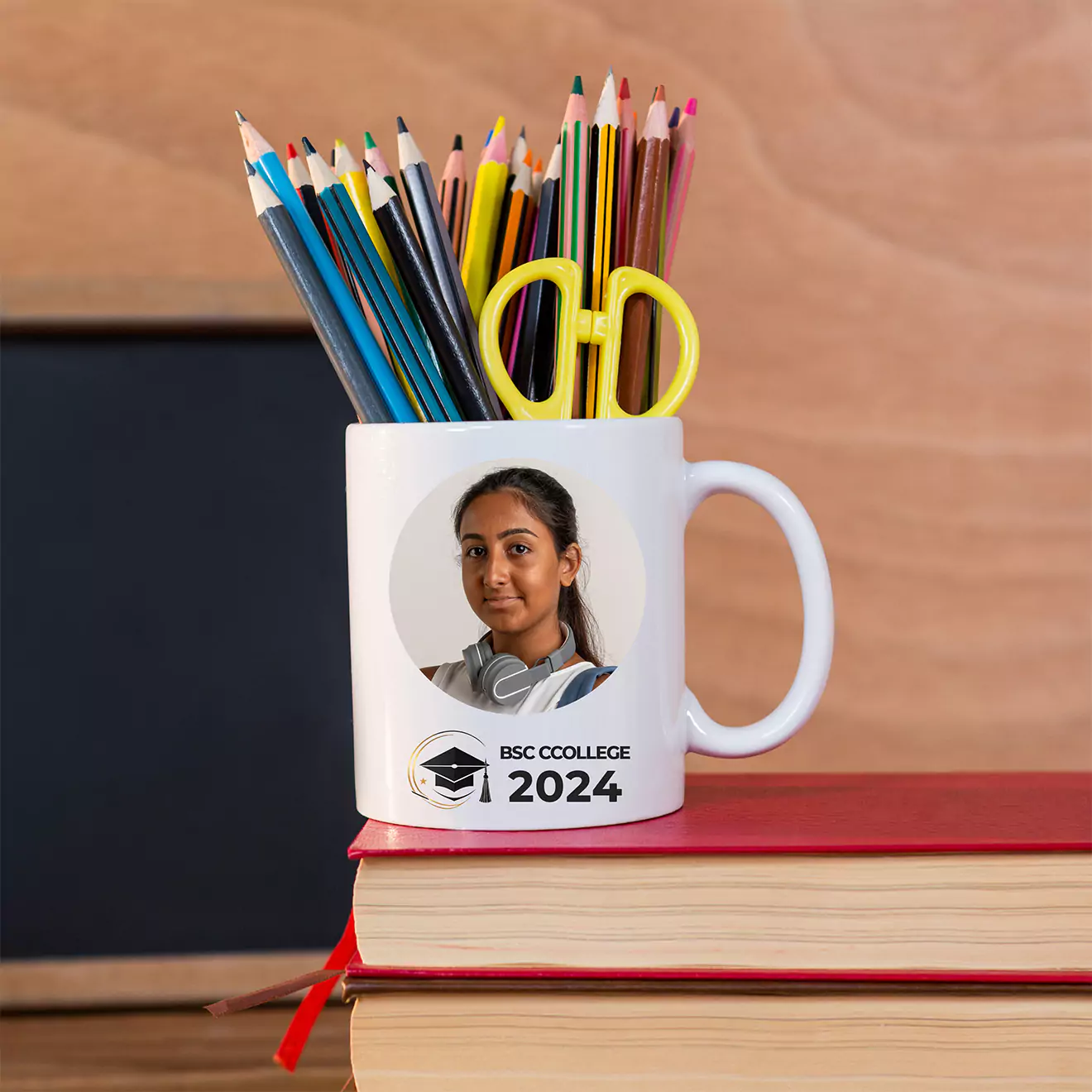 Personalised white mug featuring a student's photo and 'BSC College 2024' text, used as a pencil holder with colorful pencils and scissors, placed on top of two stacked books.