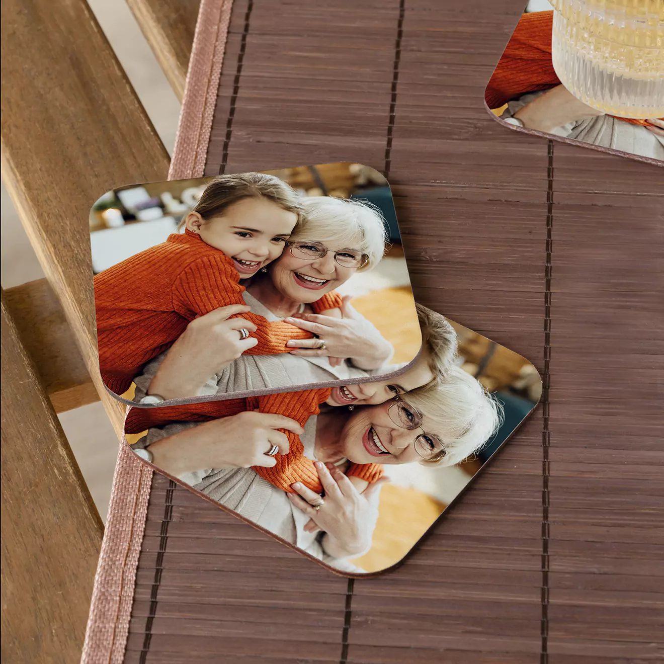 Two personalised coasters featuring a photo of a smiling elderly woman and a young girl in an orange sweater, placed on a wooden table with a woven placemat.