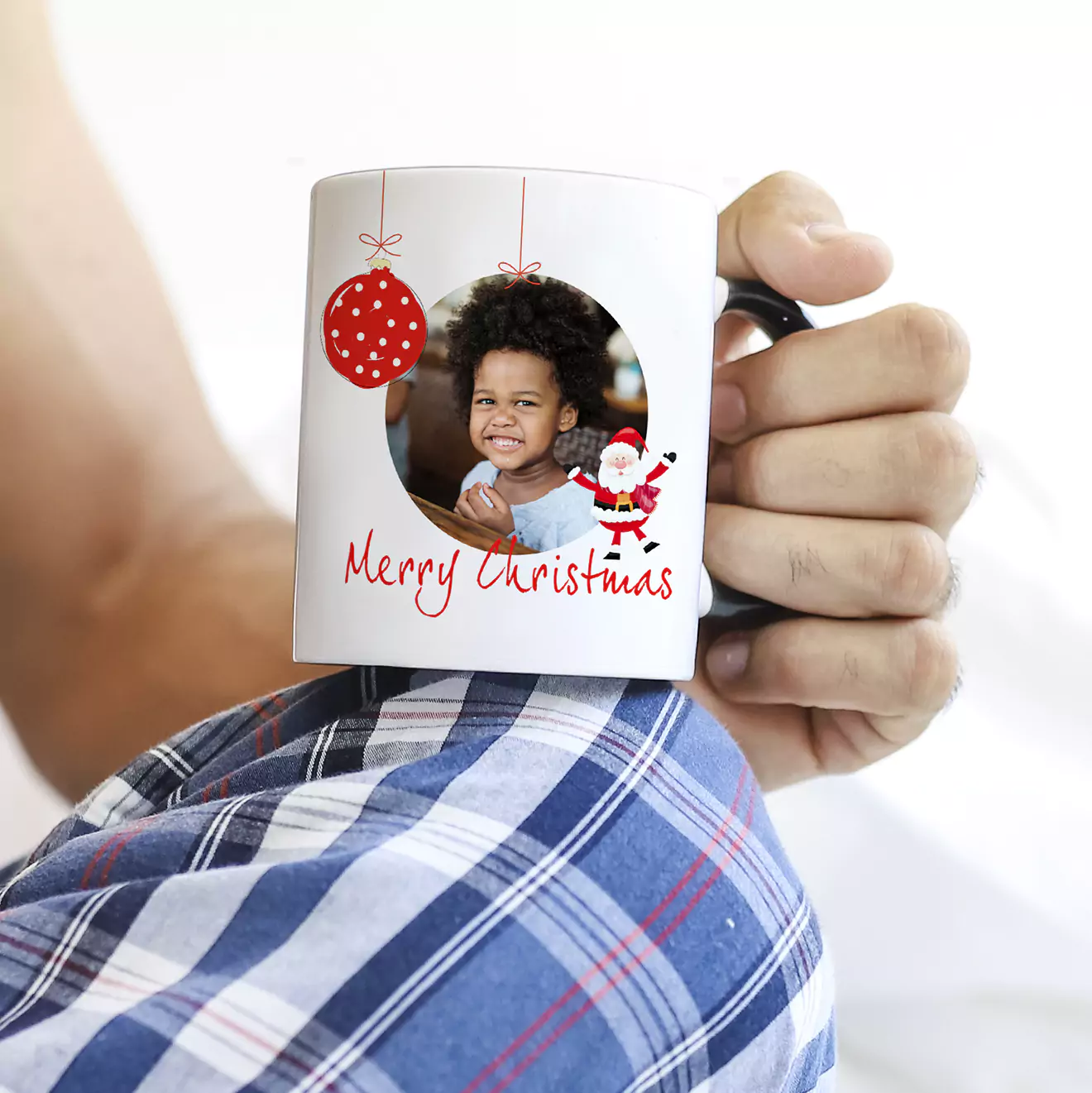 A person holding a white Christmas-themed mug featuring a photo of a smiling child, red ornaments, and the text "Merry Christmas" with a Santa Claus illustration. The person is wearing blue plaid pants.