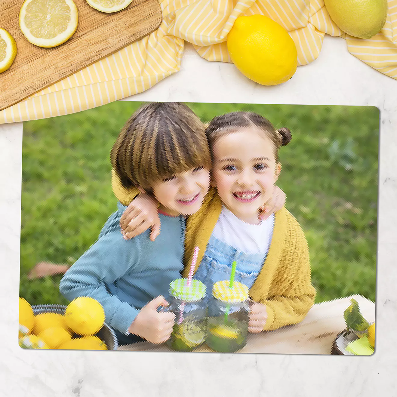 Two children smiling and hugging while holding mason jar drinks with straws, surrounded by lemons on a table. The background features a green outdoor setting, evoking a cheerful and fresh atmosphere.