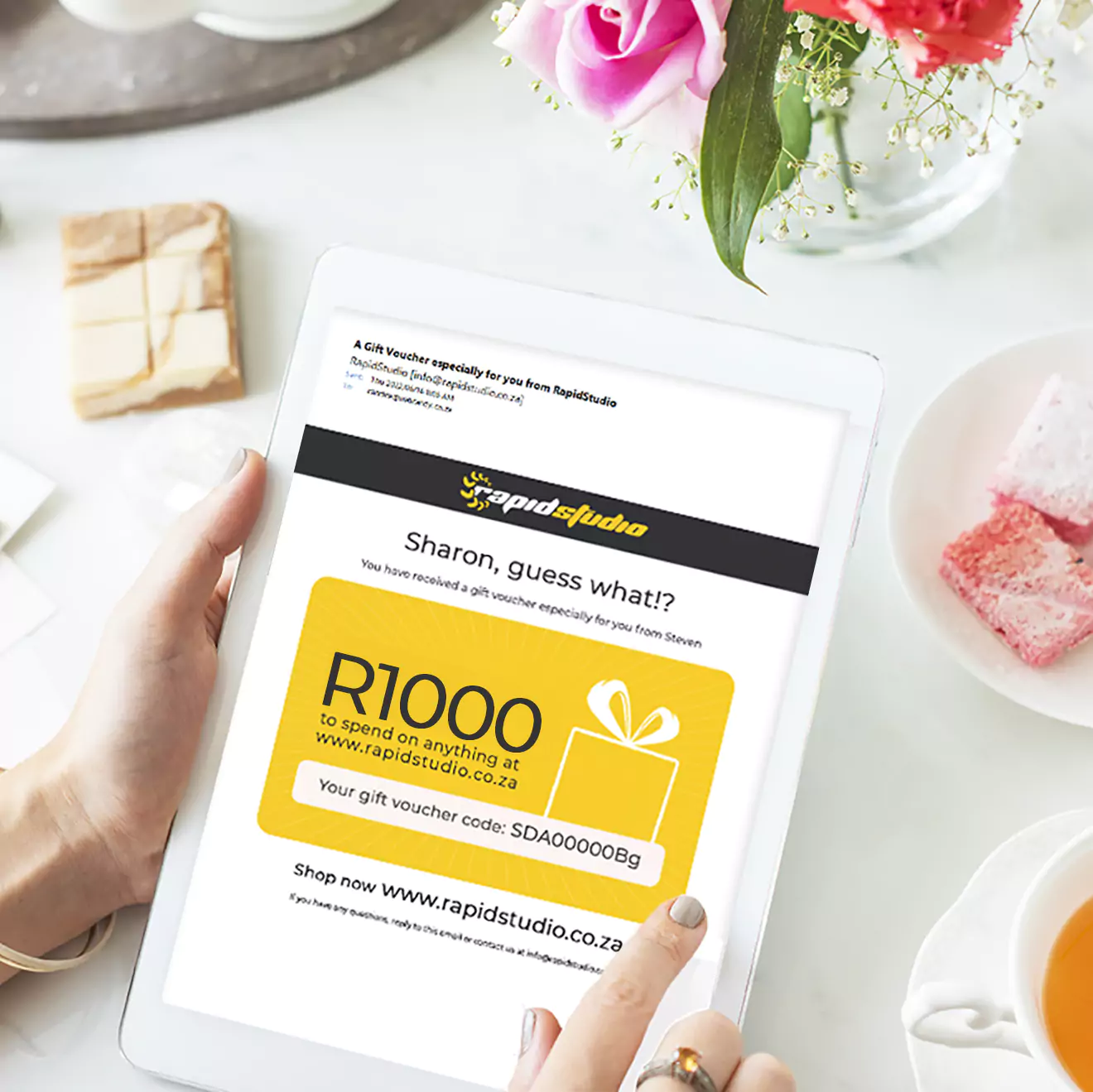 A person holding a tablet displaying a gift voucher from RapidStudio worth R1000. The tablet is on a white table with a cup of coffee, a plate with a dessert, and a vase with flowers.