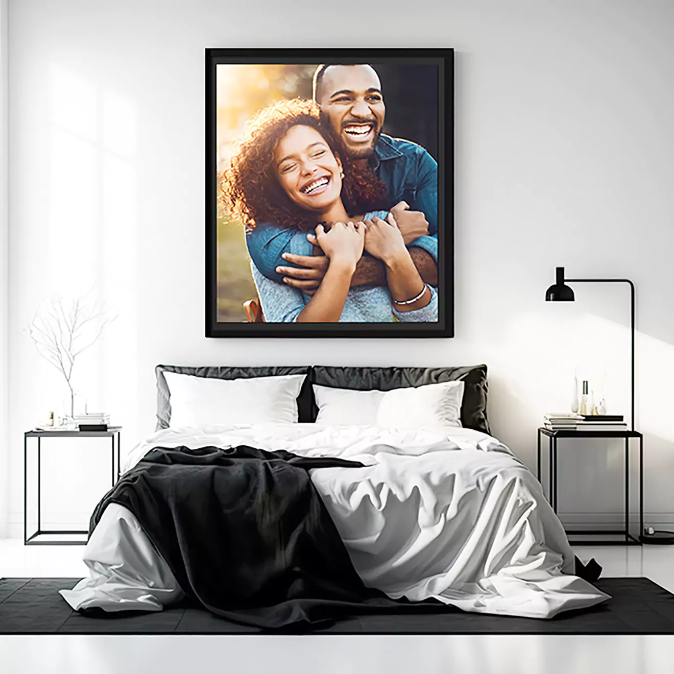 A modern bedroom with a neatly made bed featuring white and black bedding. Above the bed hangs a large framed photo of a smiling couple embracing. The room is decorated with minimalist furniture and a floor lamp.