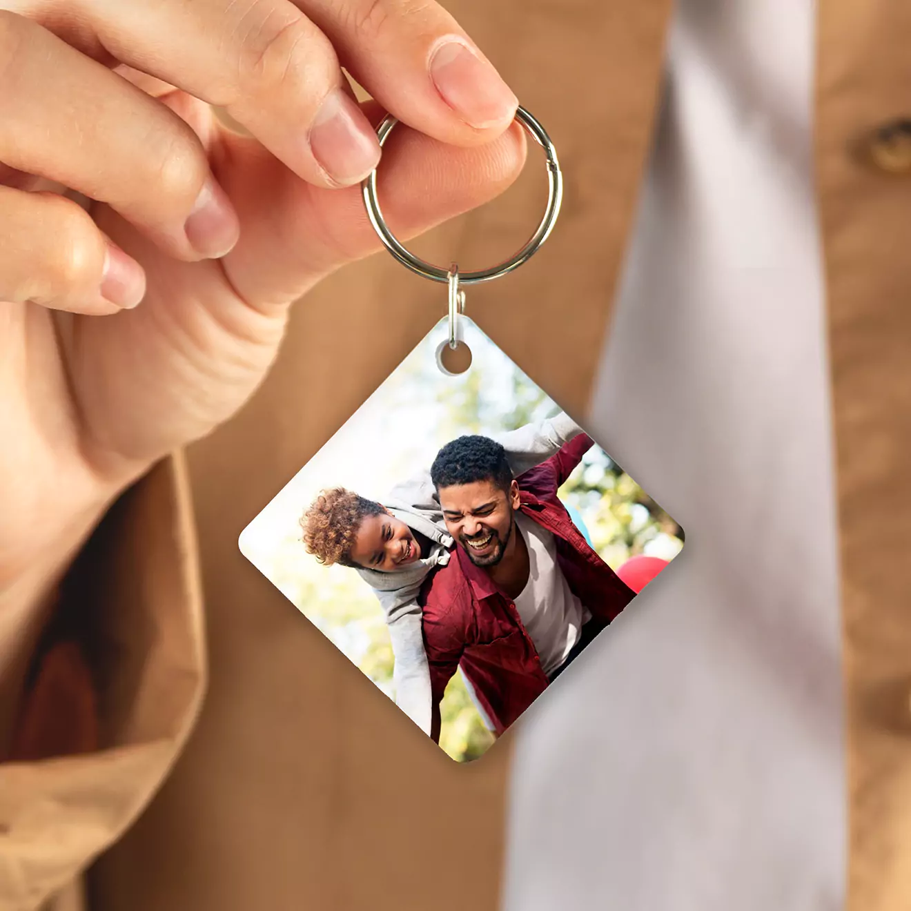 A person holding a square-shaped keychain featuring a personalised photo of two people smiling and enjoying a playful moment together. The keychain is being showcased against a blurred background.