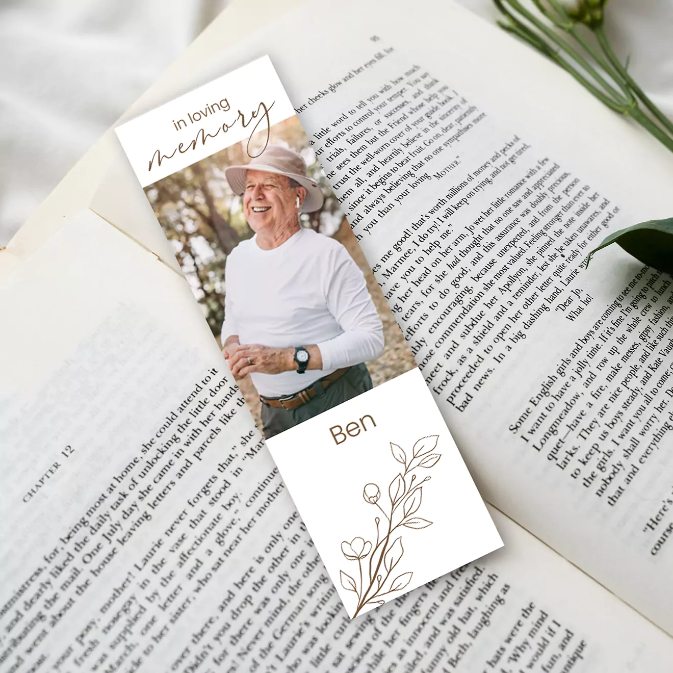 A personalised funeral memorial bookmark placed on an open book. The bookmark features a photo of an elderly person in a white shirt and hat, with the text 'In loving memory' and the name 'Ben' along with a floral illustration.