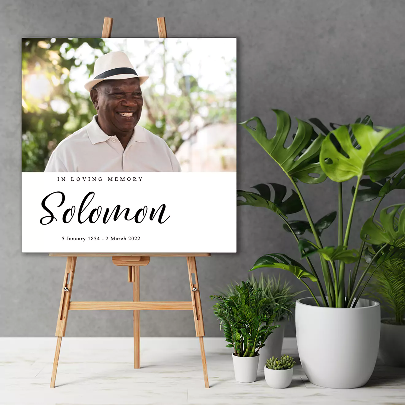 Funeral memorial photo book displayed on an easel, featuring a smiling man in a white hat with the text 'In Loving Memory Solomon, 5 January 1954 - 2 March 2022.' Surrounded by potted plants.