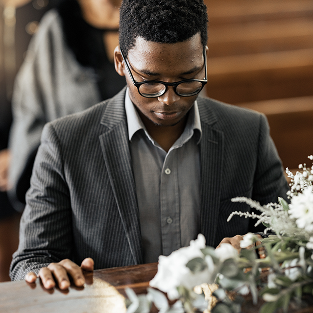 A man in a suit and glasses sits solemnly in a church pew, with a floral arrangement in the foreground, during a funeral service. The atmosphere is reflective and respectful.