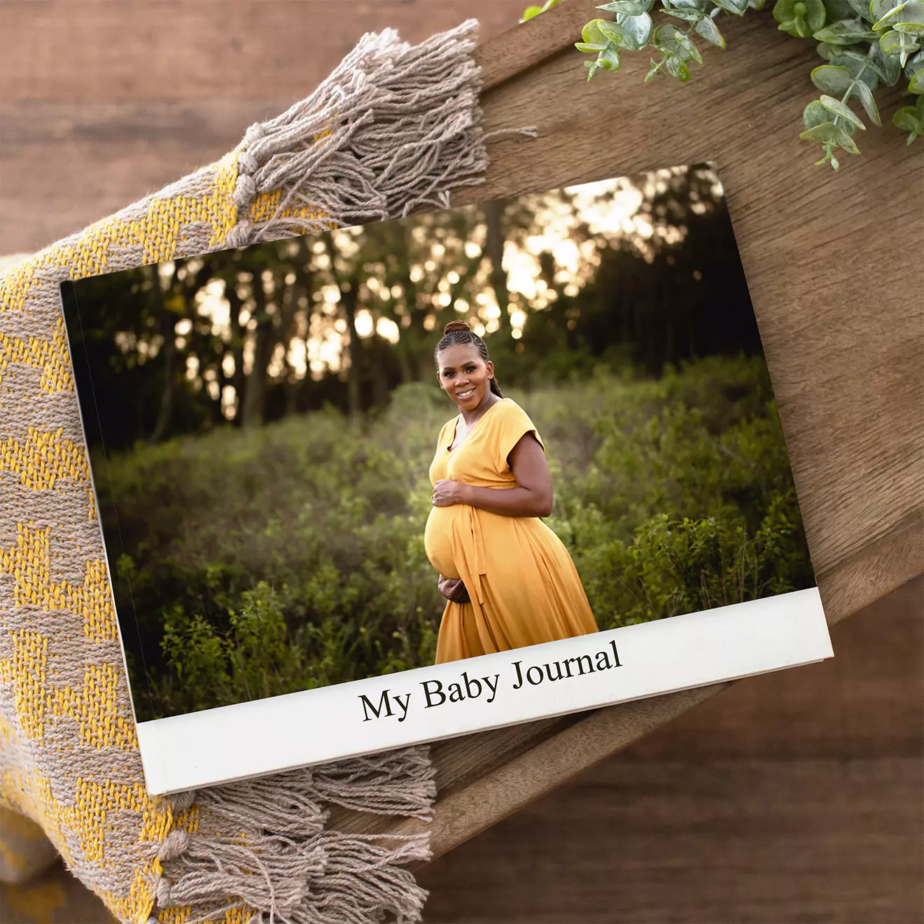 A photobook titled 'My Baby Journal' featuring a pregnant woman in a yellow dress standing in a lush green field. The photobook is placed on a wooden surface with a woven blanket.