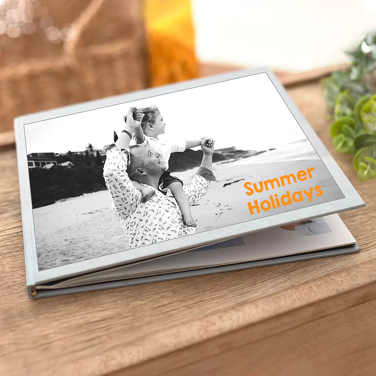 A photobook titled "Summer Holidays" featuring a black and white cover image of a person carrying a child on their shoulders at the beach. The photobook is placed on a wooden surface.