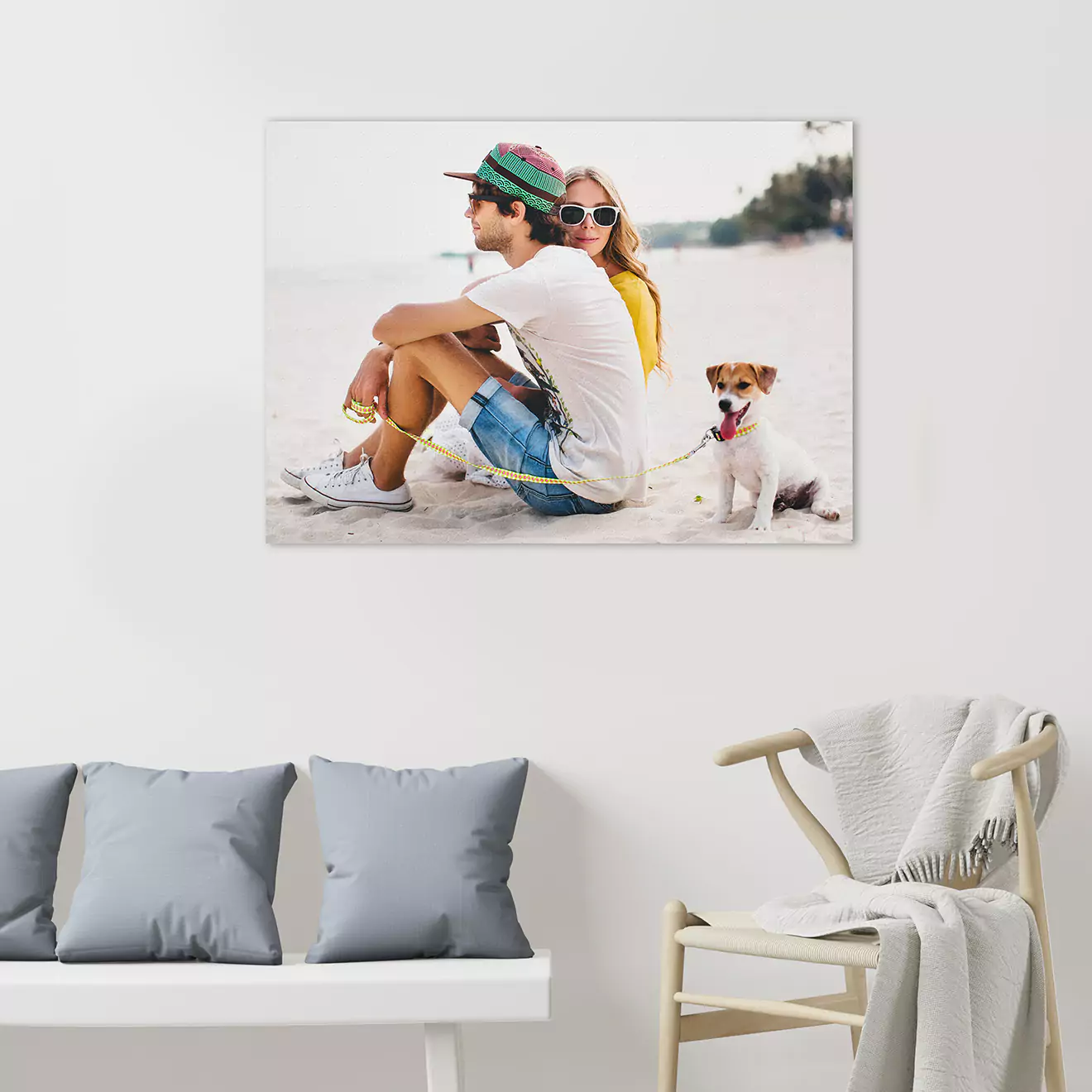 Wall-mounted photo canvas featuring a young couple sitting on a beach with a small dog. The room includes a white bench with grey cushions and a white chair draped with a blanket.