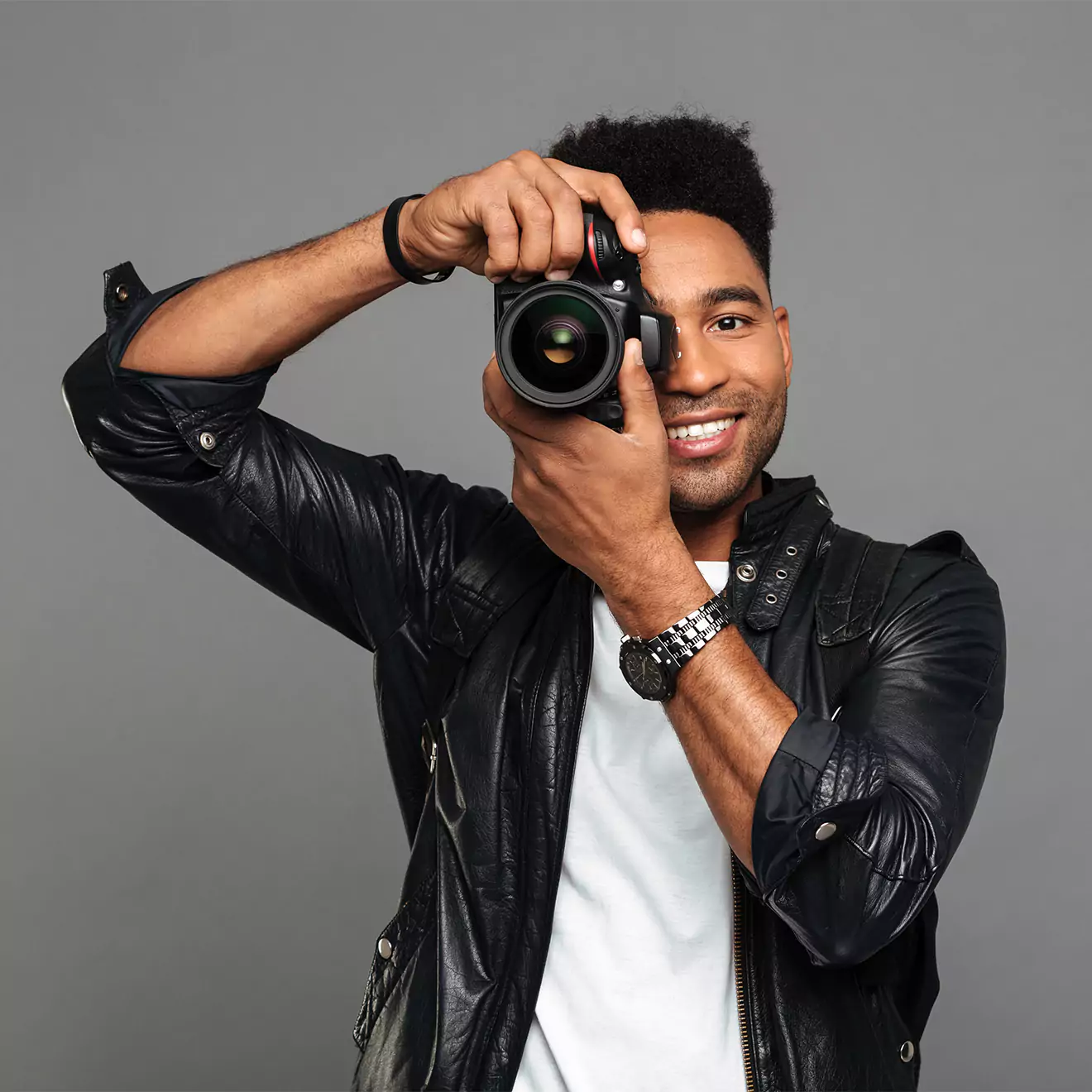 A smiling person wearing a black leather jacket and white shirt holds a camera up to their face, ready to take a photo. The background is a plain grey, highlighting the subject.