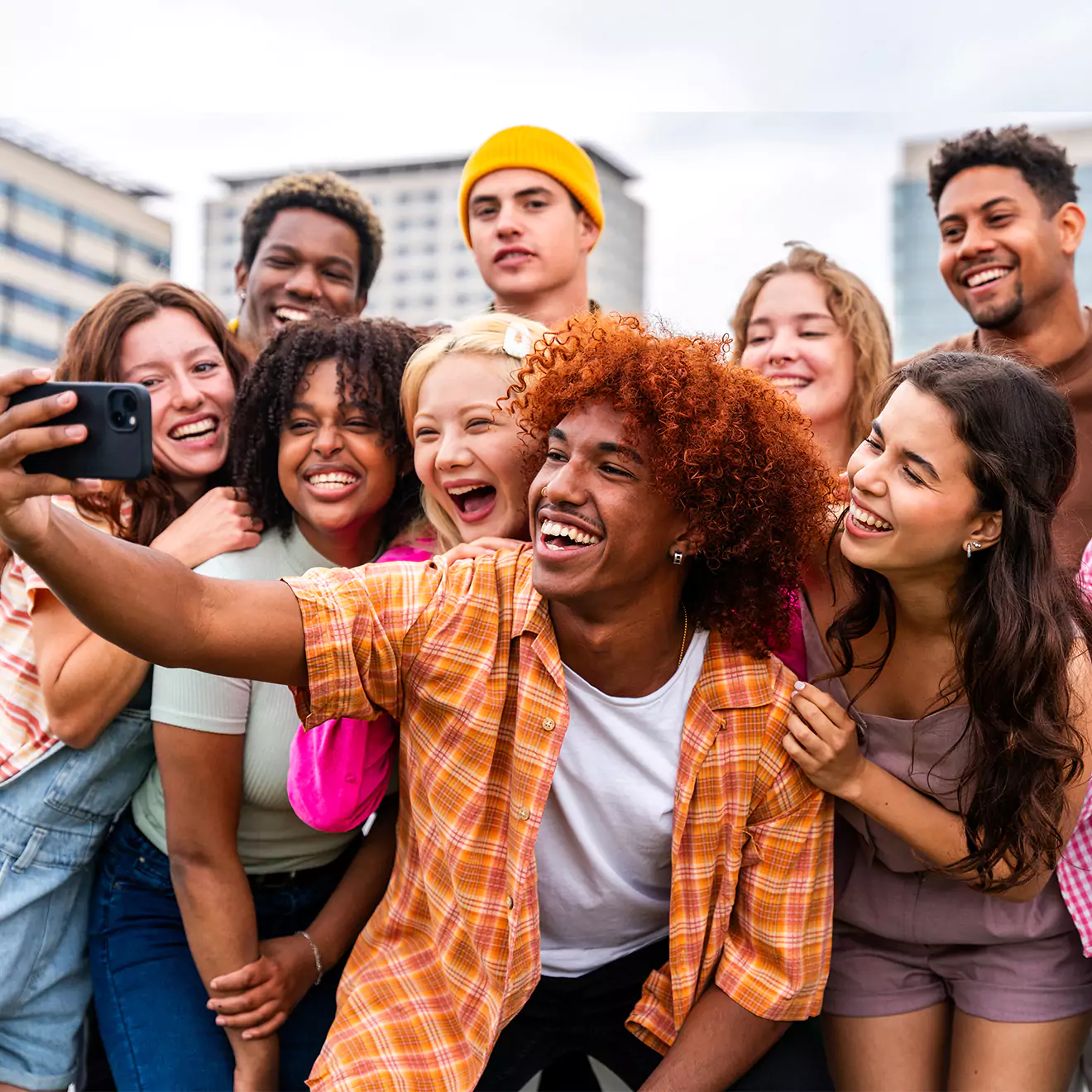 A group of diverse young adults smiling and taking a selfie outdoors, with buildings in the background. They appear to be enjoying a casual, joyful moment together.