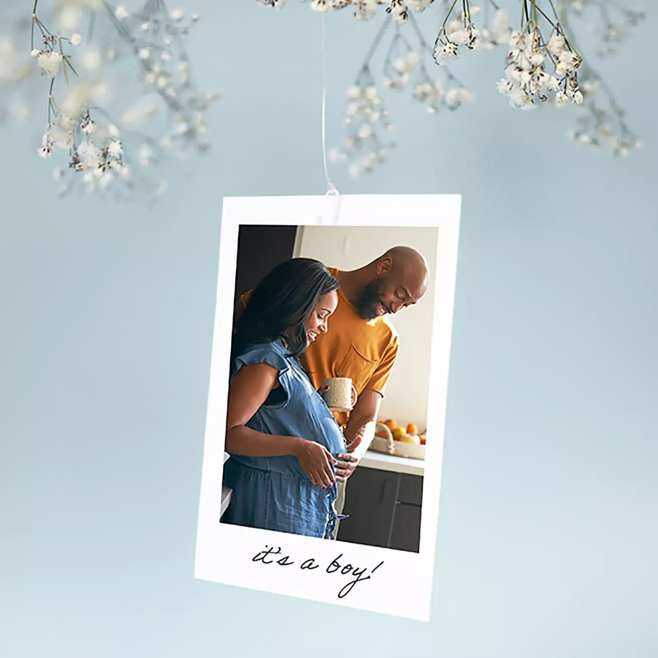 Mini print featuring a photo of a smiling couple, with the woman holding her pregnant belly and the man standing beside her. The print is hanging from a branch with white flowers and has the text 'it's a boy!' at the bottom.