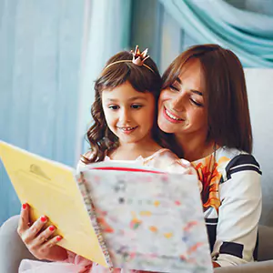 A mother and daughter share a joyful moment while reading a colorful photobook together, perfect for Mother's Day celebrations. The mother smiles warmly as the daughter, wearing a crown, looks at the book.