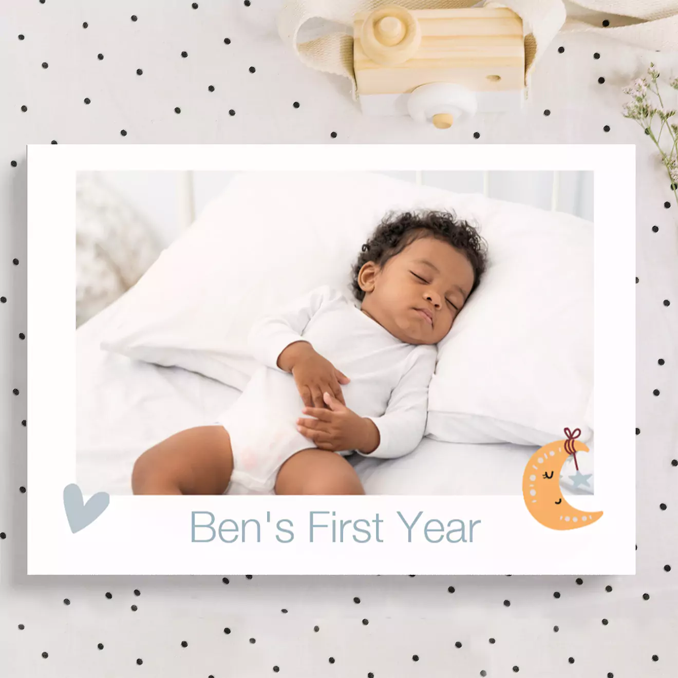 Personalised baby gift featuring a photo of a sleeping baby on a white pillow with the text 'Ben's First Year' below. The design includes a small heart and moon illustration.