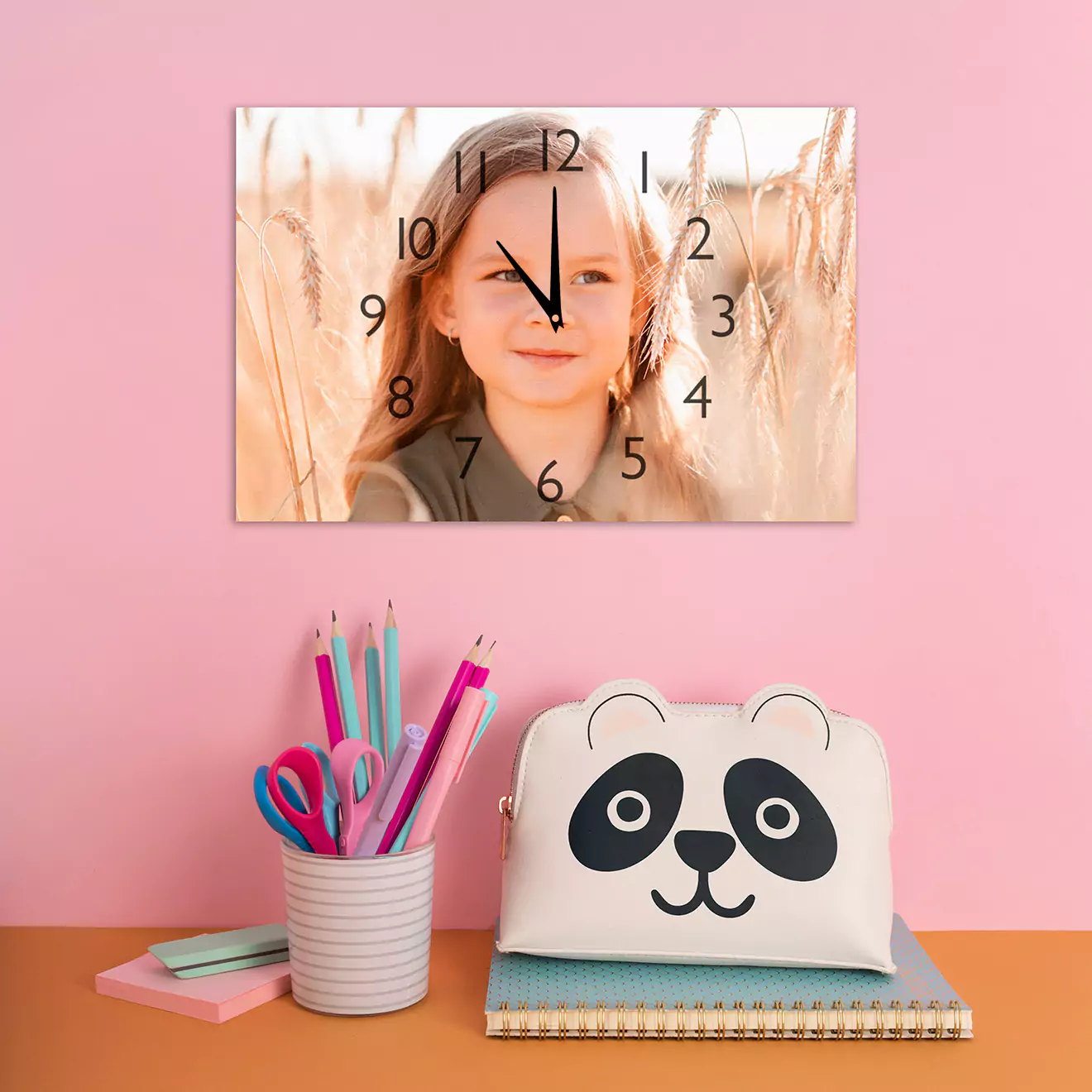 Personalised baby gift featuring a custom photo wall clock with a child's image, displayed above a desk with a panda-themed pencil case, notebooks, and a cup holding colourful pens and pencils.