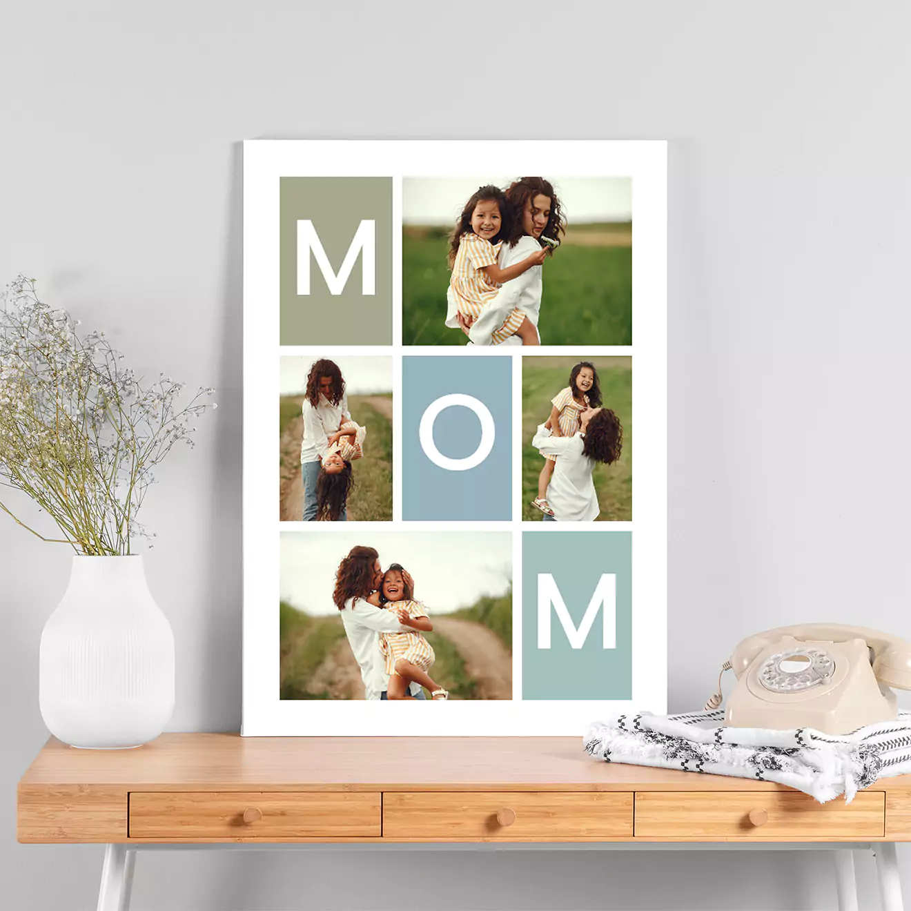 A personalised photo collage print featuring the word "MOM" with various images of a mother and child in outdoor settings. The print is displayed on a wooden table next to a vase with flowers and a folded blanket.