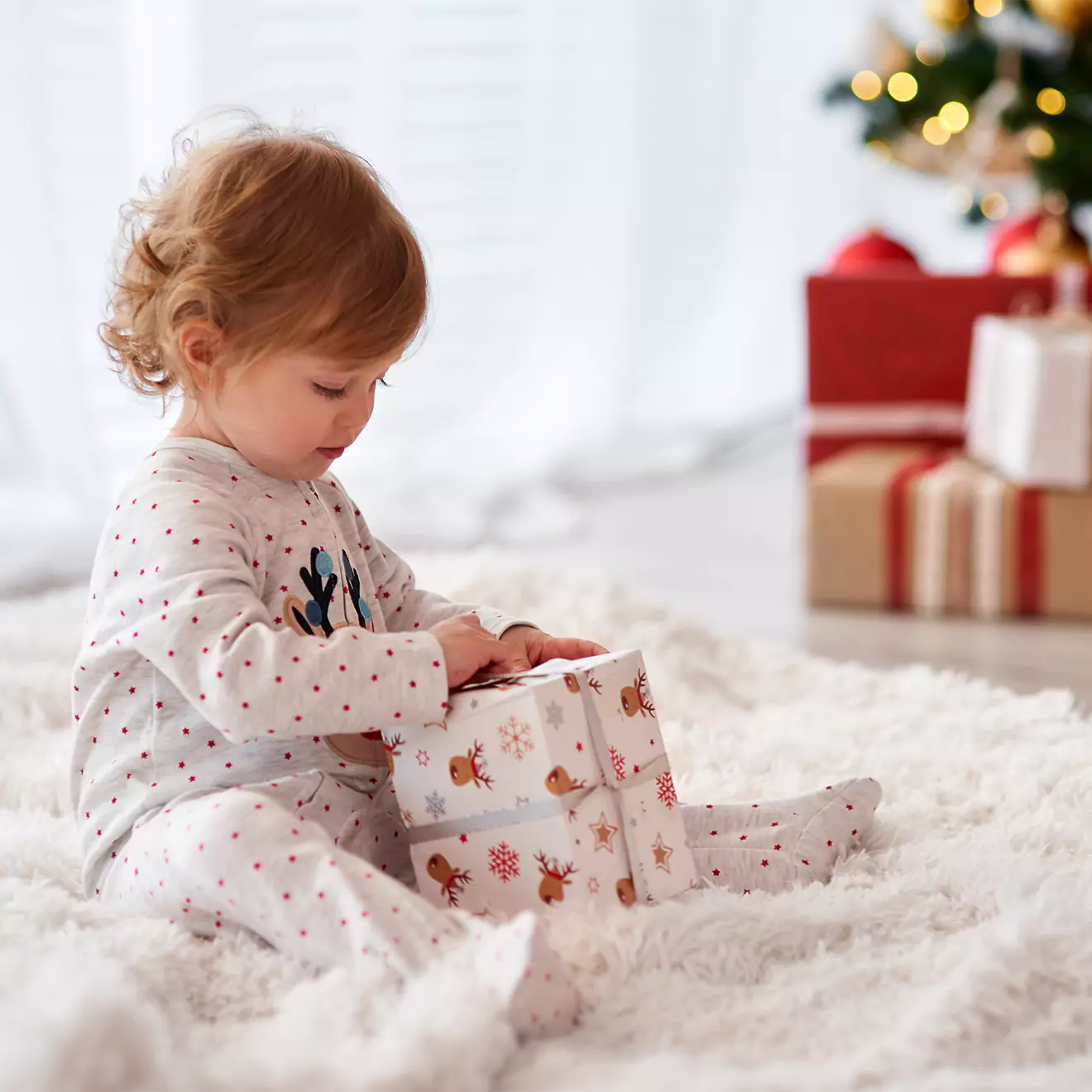 A young child in festive pajamas sits on a fluffy white rug, eagerly opening a wrapped gift. In the background, there are more wrapped presents and a decorated Christmas tree.