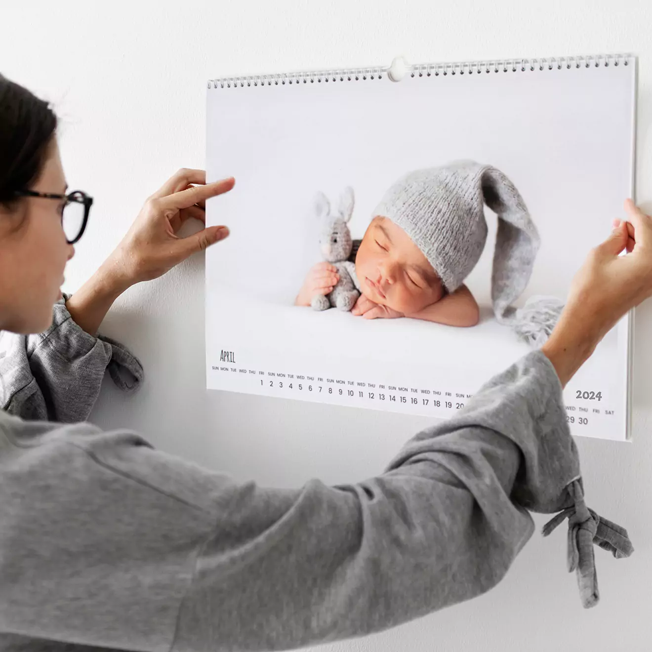 A person is hanging a personalised wall calendar featuring a sleeping baby wearing a grey knitted hat and cuddling a stuffed animal. The calendar is open to April 2024.