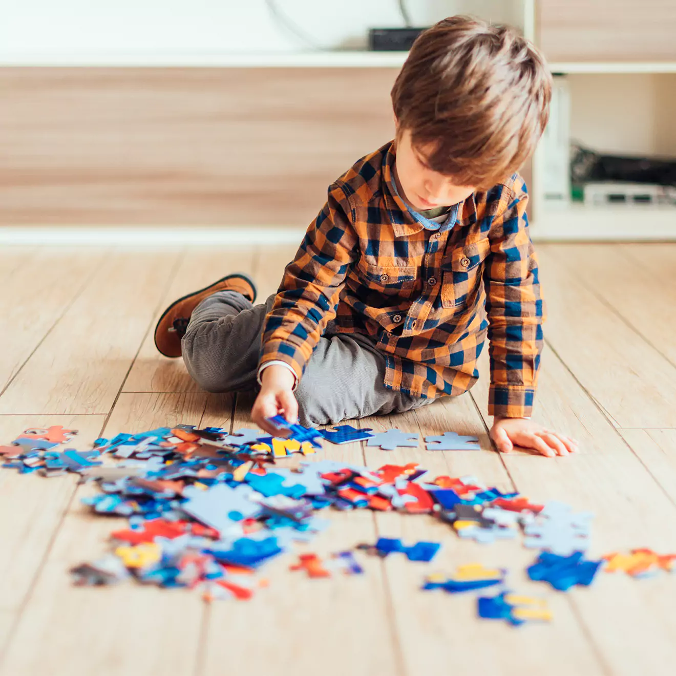 A young child in a plaid shirt and gray pants is sitting on a wooden floor, focused on assembling a colorful jigsaw puzzle. The background features a white shelf with various items.