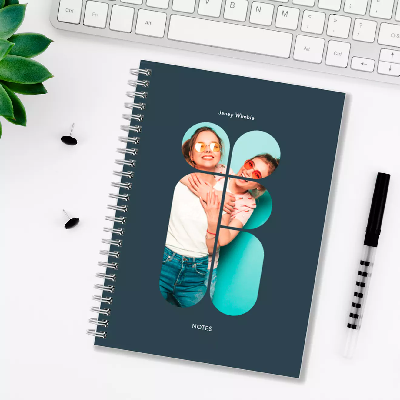 Personalised softcover notebook with a spiral binding, featuring a custom photo of a smiling couple on the cover. The notebook is placed on a white desk with a keyboard, pen, and plant.