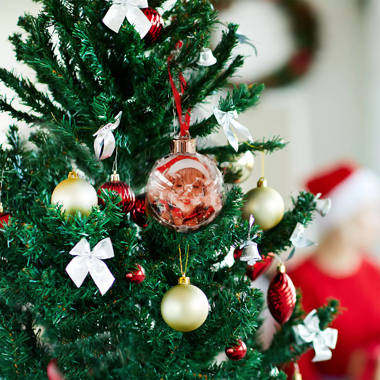 A decorated Christmas tree adorned with various ornaments, including gold and red baubles, white bows, and a clear bauble with a festive design. A blurred figure in a Santa hat is visible in the background.