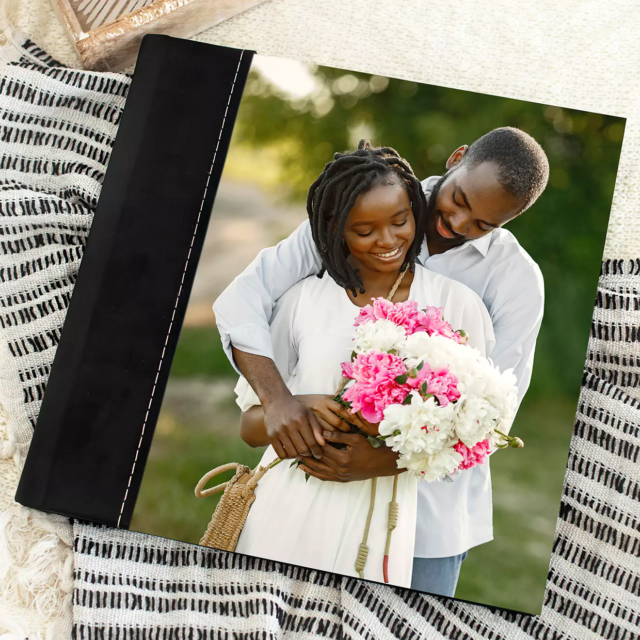 A photo Album with a black leather spine and a cover image of a smiling couple embracing, with the woman holding a bouquet of pink and white flowers. The Designer Album is placed on a striped fabric background.