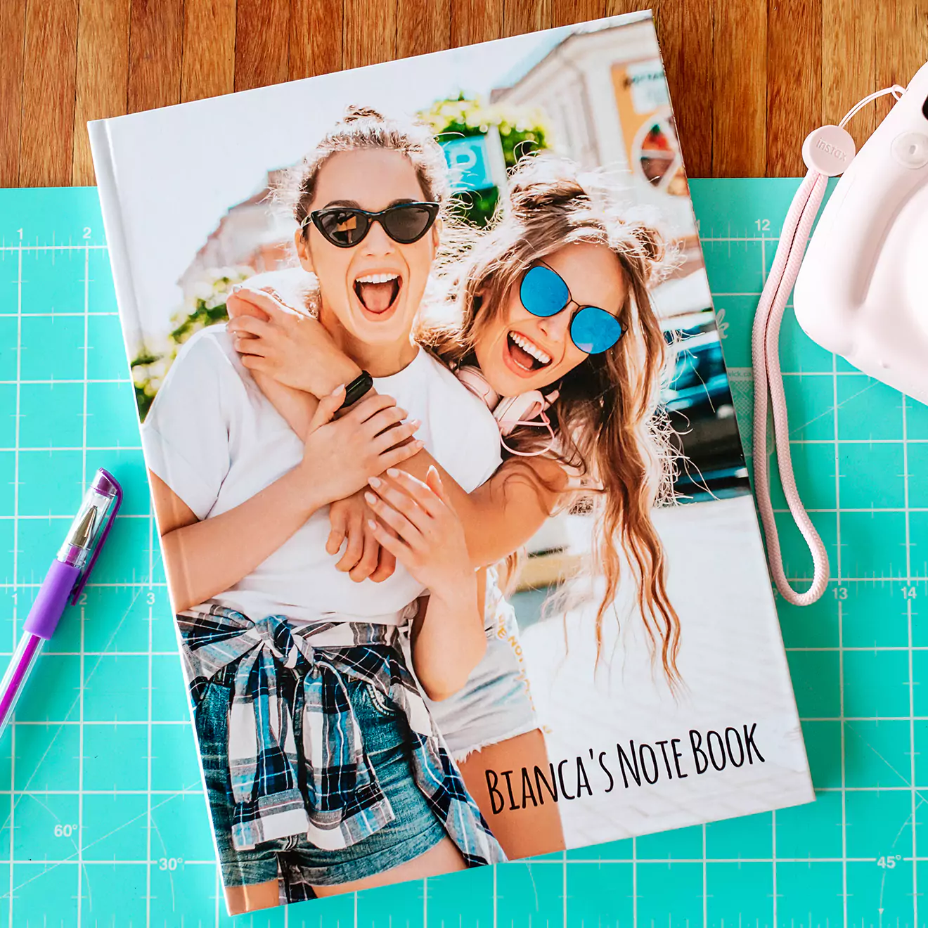 A personalised hardcover note book titled "Bianca's Note Book" featuring a cover photo of two smiling women wearing sunglasses. The photobook is placed on a blue cutting mat with a purple pen and a white camera nearby.