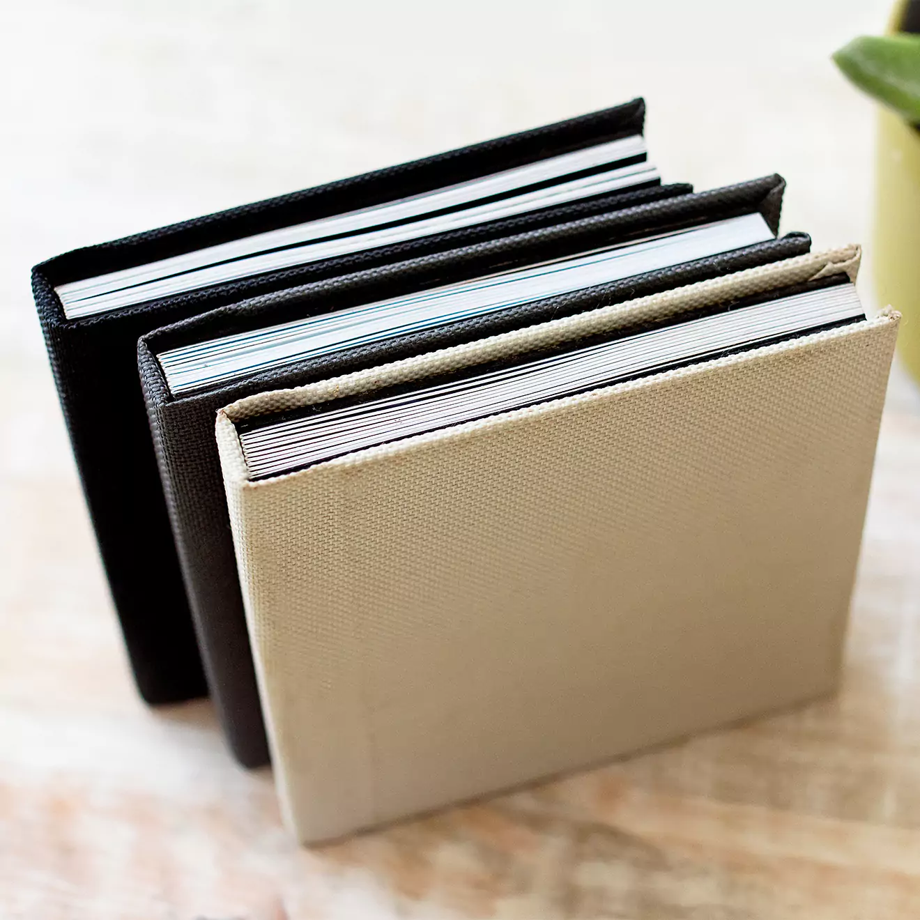 Three hardcover photobooks with textured covers in black, dark brown, and beige are neatly stacked on a light-colored surface. The photobooks are positioned upright, showcasing their spines and pages.