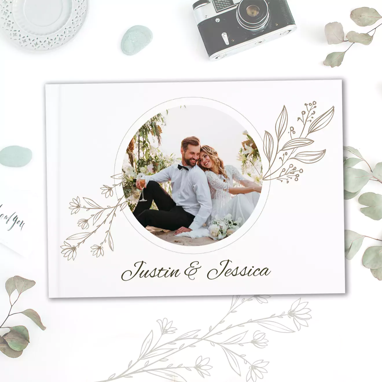 Elegant photobook cover featuring a circular photo of a couple sitting outdoors, surrounded by delicate floral illustrations. The names 'Justin & Jessica' are written below the photo.