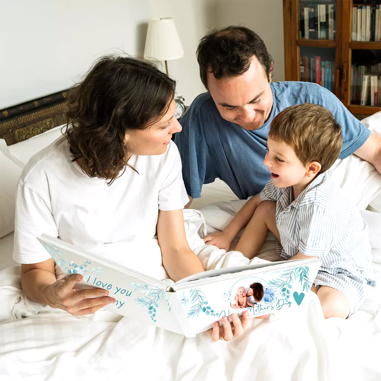 A family of three, including a mother, father, and young child, sits on a bed looking at a personalised photobook together. The photobook features a cover with a photo and the text "I love you.